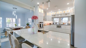 Una cocina moderna con armarios blancos, una isla de mármol, iluminación colgante y un comedor con mesa y sillas.