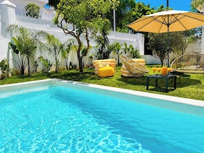 Ein Schwimmbecken mit klarem, blauem Wasser, umgeben von Rasenflächen, Pflanzen und Gartenmöbeln.