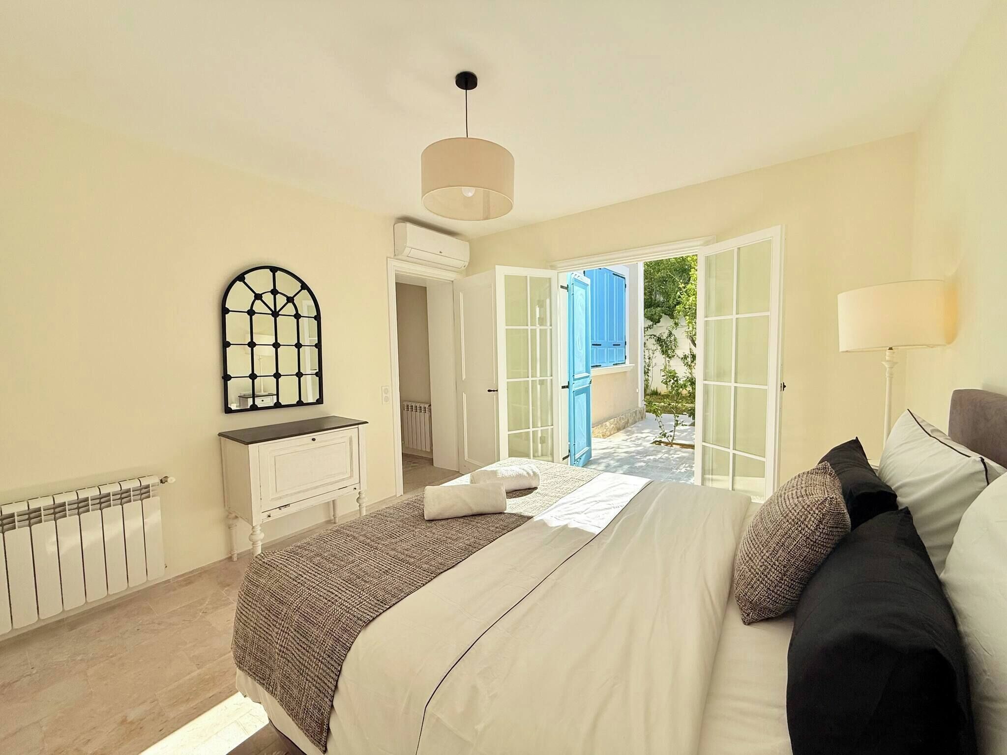 A bedroom with a bed, bedside table, lamp, and a view through an open door to a courtyard.