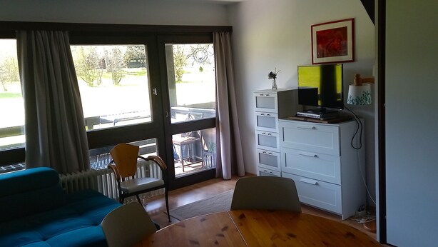 A room with a blue sofa, a wooden dining table, a TV on a cabinet, and a view through large windows. - Vacation apartment Iris 5 (Siegsdorf)