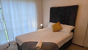 Una cama bien hecha con cabecera, lámpara de noche y ventana con cortinas.