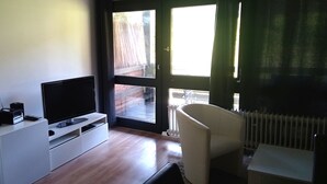 A modern living room with a flat-screen TV, a white entertainment unit, a white chair, and a large window with a view of a wooden deck. - Vacation apartment Glock 6 (Siegsdorf)