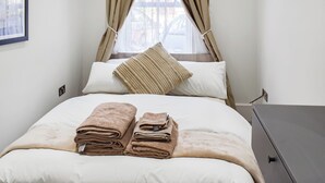 A neatly made bed with folded towels, a pillow, and a window with curtains. - Apartment 2 - UK51647 (Maldon)