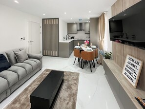 A modern living room with a sofa, a dining table set for six, a kitchen in the background, and a mounted TV. - Apartment 4 - UK51649 (Maldon)