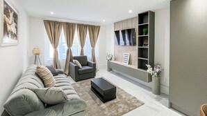 A modern living room with a grey sofa, a black ottoman, and a grey armchair. There is a bookshelf with decorative items and a sign.