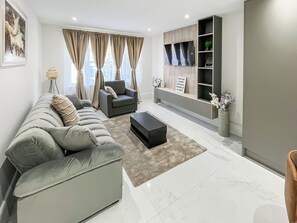 A modern living room with a grey sofa, a black ottoman, and a grey armchair. There is a bookshelf with decorative items and a sign. - Apartment 1 - UK51646 (Maldon)