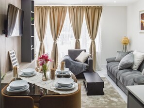 A modern living room with a dining area, grey sofas, beige curtains, and a flat-screen TV mounted on the wall. - Apartment 1 - UK51646 (Maldon)