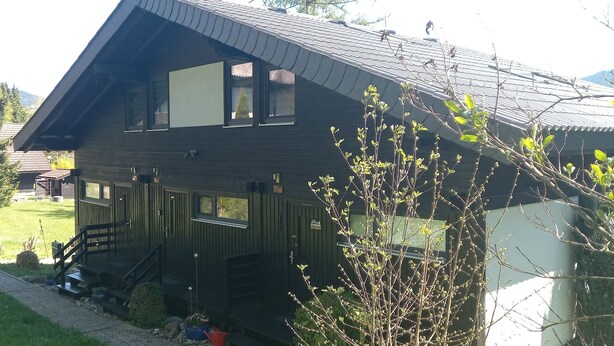 A two-story wooden house with a porch, a small garden, and a mountain backdrop.