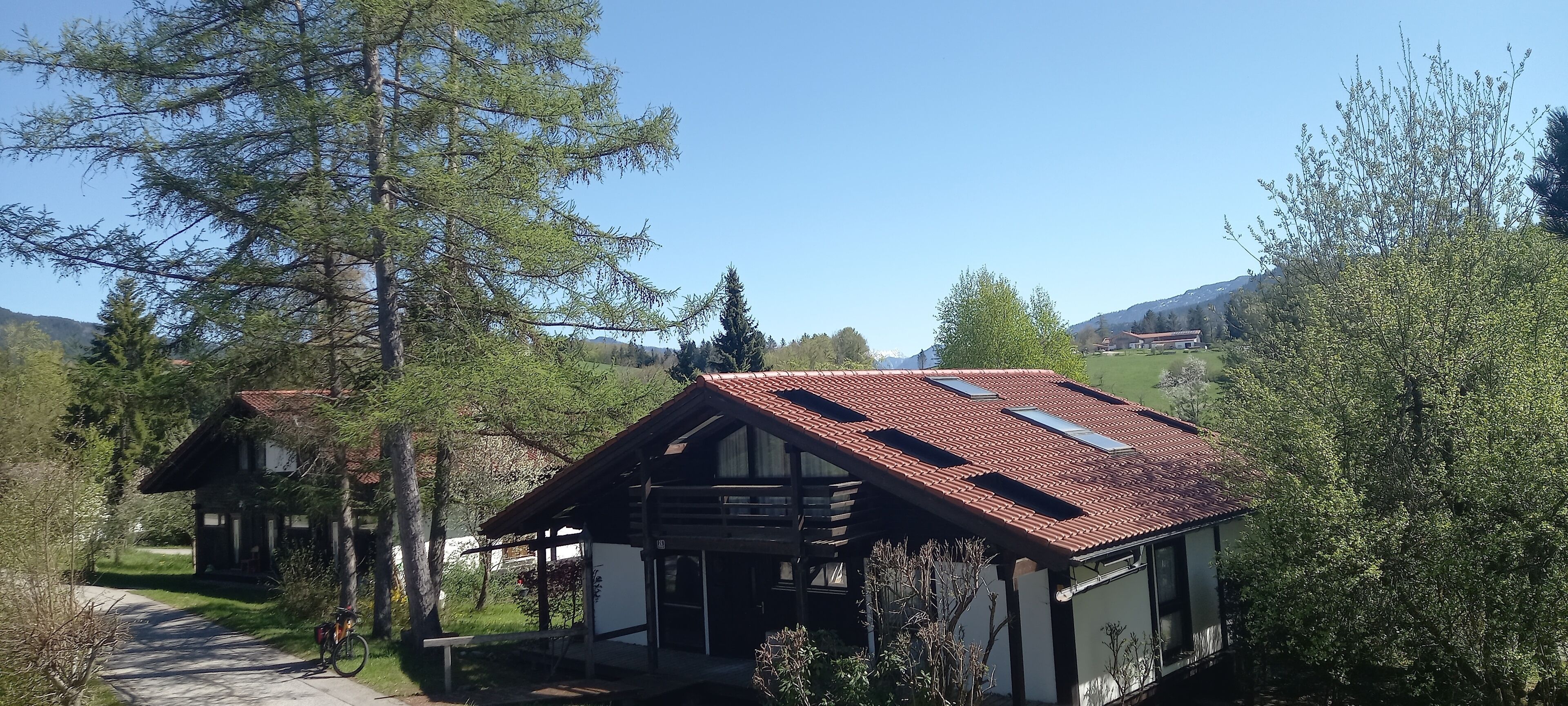 Ein traditionelles Chalet mit geneigtem Dach, einer Holzterrasse und Blick auf den umliegenden Wald und die Berge.