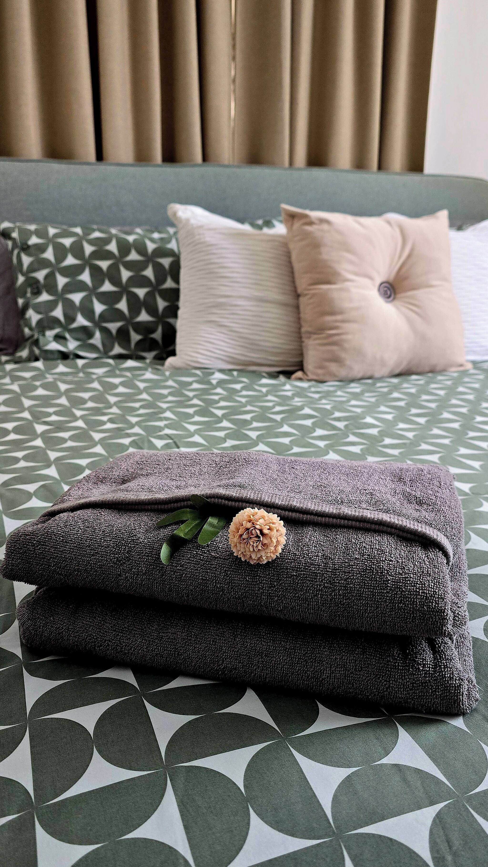 A neatly made bed with a geometric patterned bedspread, two pillows, and a folded towel with a decorative flower.