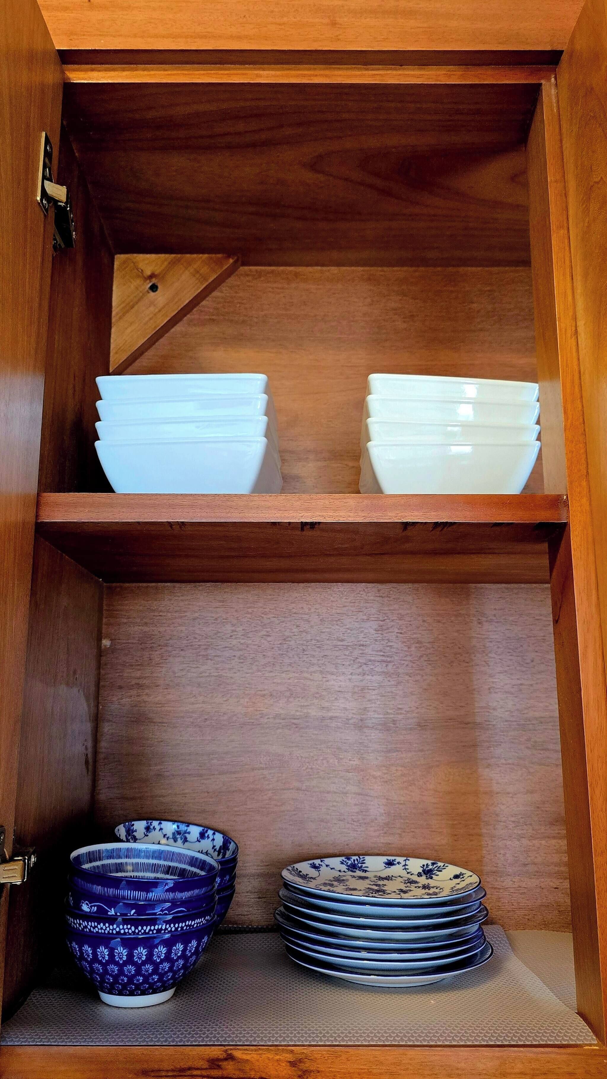 Un gabinete de madera con dos estantes; el estante superior contiene cuencos blancos, y el estante inferior tiene una pila de cuencos con motivos azules y blancos.