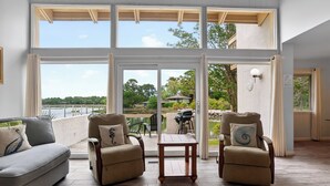 Une pièce comprenant un canapé, deux fauteuils, une petite table et de grandes fenêtres donnant sur un lac, avec un espace aménagé à l’extérieur.