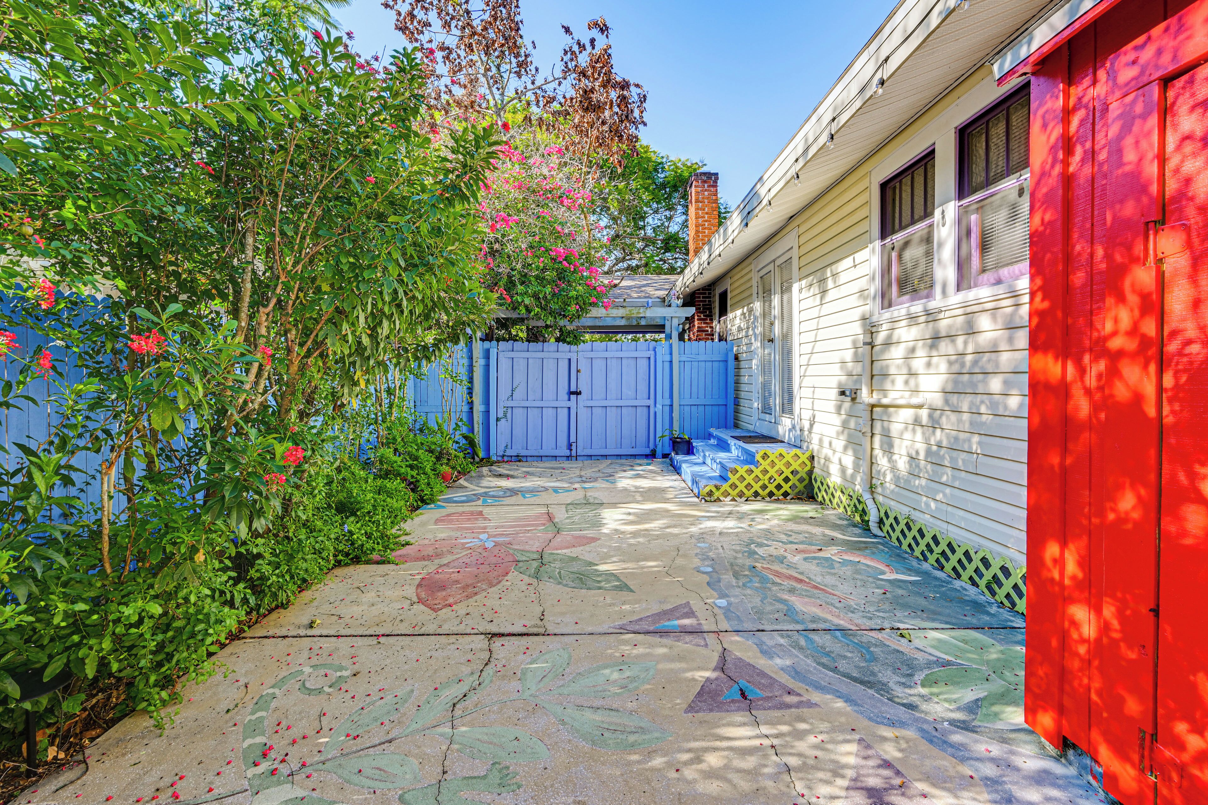 Ein Garten mit Wohnhäusern, einem blauen Tor, einer roten Gartenhütte und einer Beton-Terrasse mit farbenfrohen Kunstwerken.