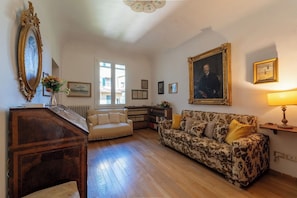 A room with a wooden floor, a sofa, a desk, a large framed portrait, and a window. - Casa dello Scrittore (Florence)