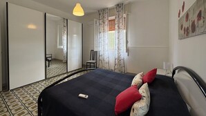 Ein Schlafzimmer mit Bett, Sessel, Schreibtisch, Spiegel und einem Fenster mit Vorhängen.