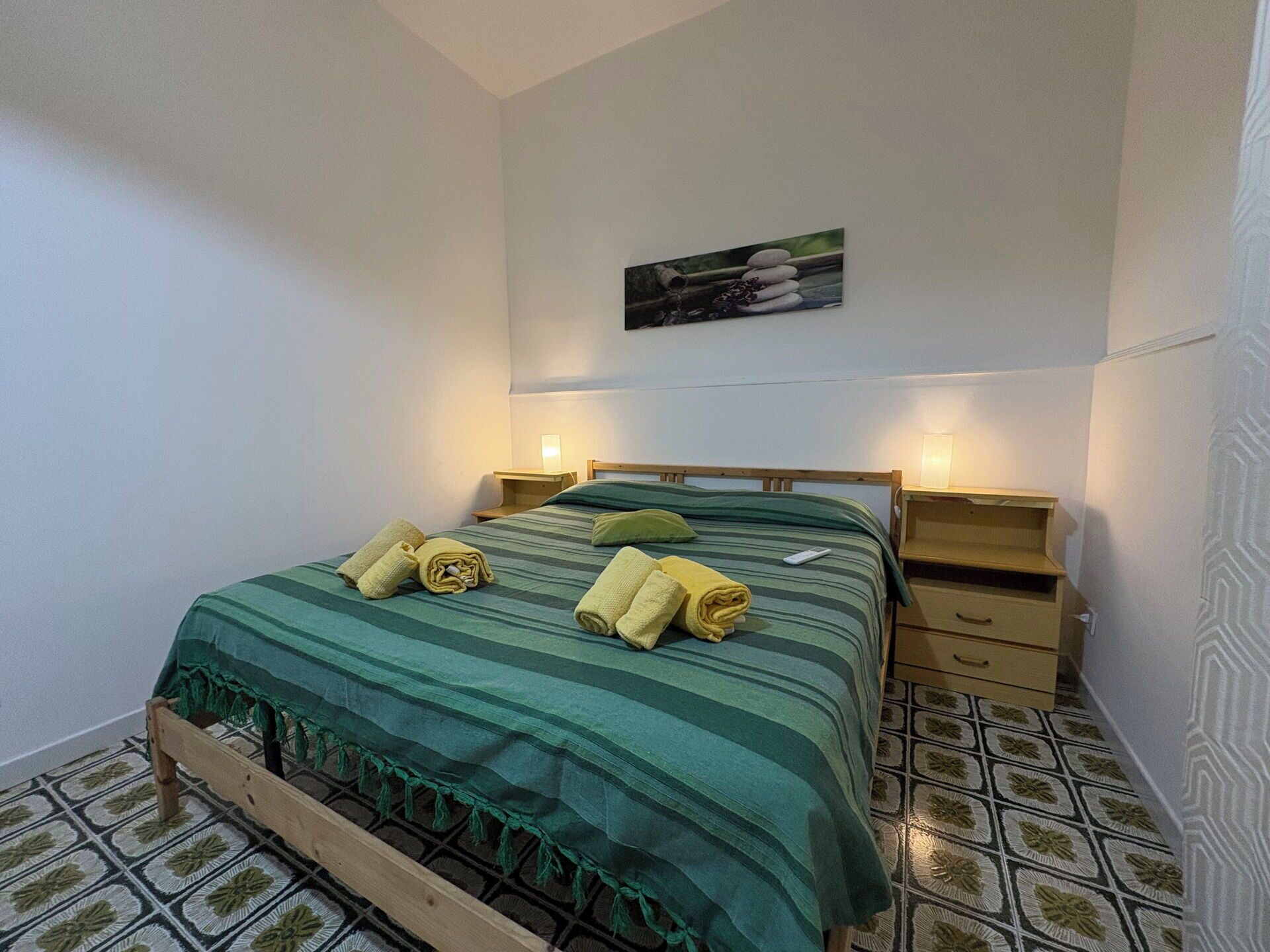 A single-bed room with a wooden frame, green striped bedding, two bedside tables with lamps, and a wall-mounted picture.