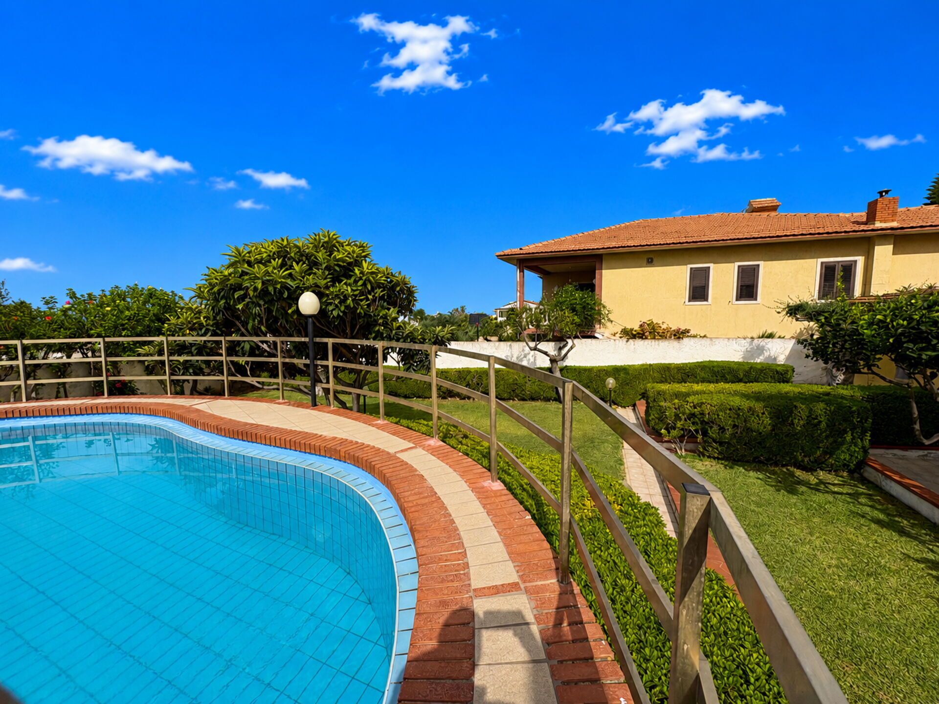 Piscina com borda azulejada, rodeada por um deck de tijolos e metal, com uma casa ao fundo.