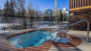 Un bain à remous doté d’une balustrade en métal, entouré d’une bordure en briques, se dressant au milieu d’un paysage enneigé avec des montagnes en arrière-plan.