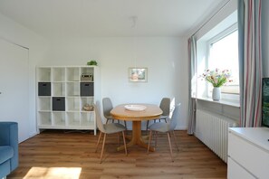 Ein Zimmer mit einem runden Holztisch, vier grauen Stühlen, einem weißen Schrank, einem weißen Regal und einem Fenster mit Vorhängen.
