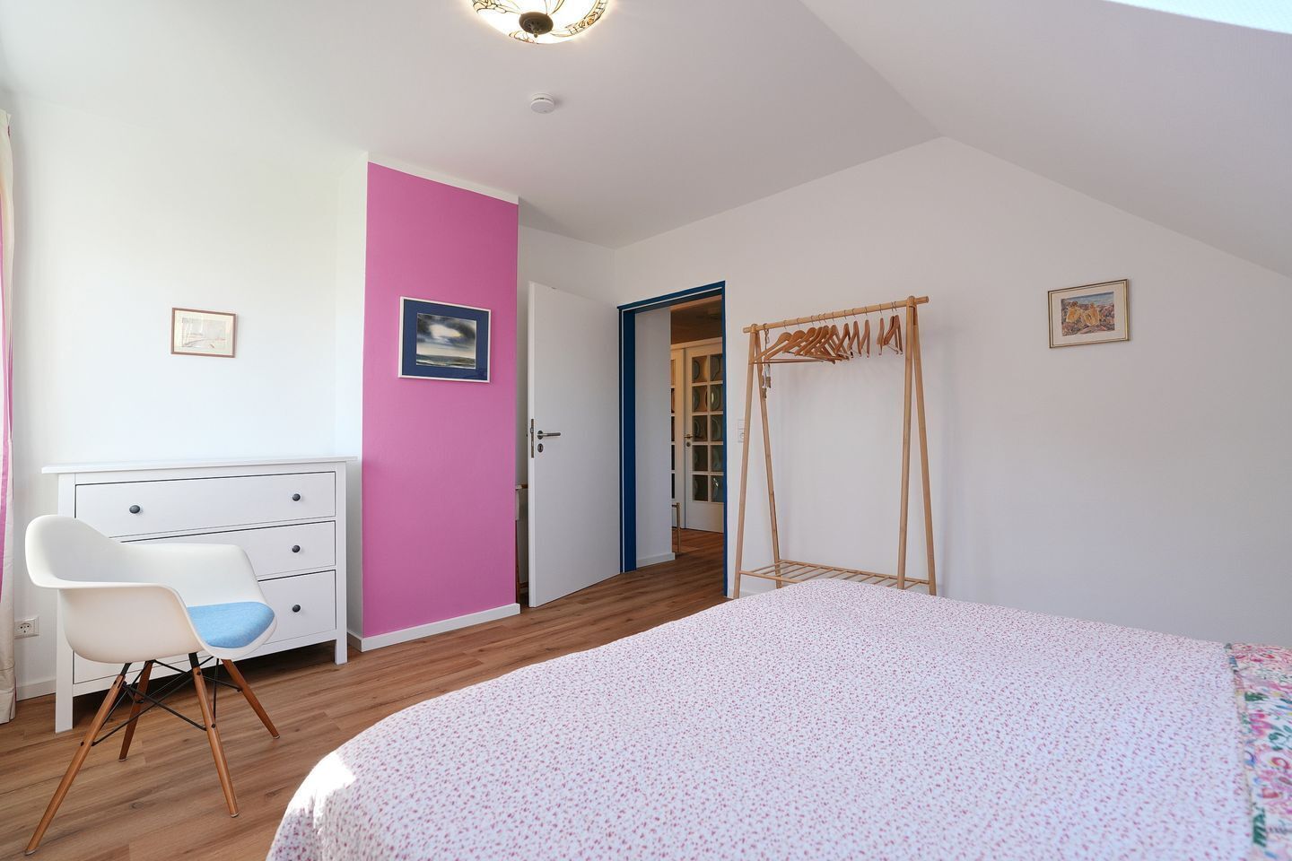 A bedroom with a bed, a white dresser, a pink door, a wooden wardrobe, and a door leading to a hallway.