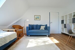 Ein Zimmer mit einem blauen Sofa, einem Korbstuhl, einem weißen Schreibtisch und einem Bücherregal.