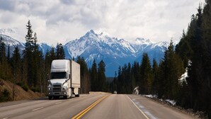 Una strada fiancheggiata da montagne innevate e una fitta foresta, con un camion di grandi dimensioni e un veicolo più piccolo.