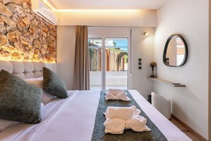 A hotel room with a stone wall, a bed with pillows, a bedside table with a vase, and a view through a glass door. - Two Bedroom Villa with Private Pool (Rhodes)