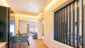 A modern interior with a dining area, kitchen, and a view through a window with vertical blinds. - Two Bedroom Villa with Private Pool (Rhodes)