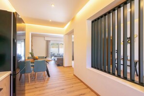 A modern interior with a dining area, kitchen, and a view through a window with vertical blinds. - Two Bedroom Villa with Private Pool (Rhodes)