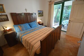 Ein Schlafzimmer mit Bett, Nachttischen, einem Kruzifix an der Wand und Blick durch eine Tür.