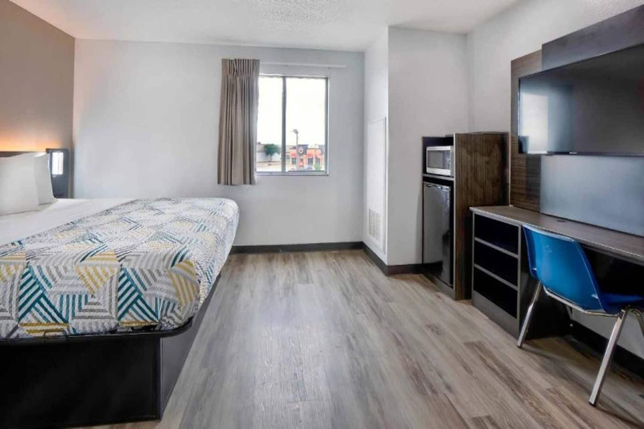 A hotel room with a bed, a desk, a chair, a TV, a window with curtains, and a view of buildings outside.