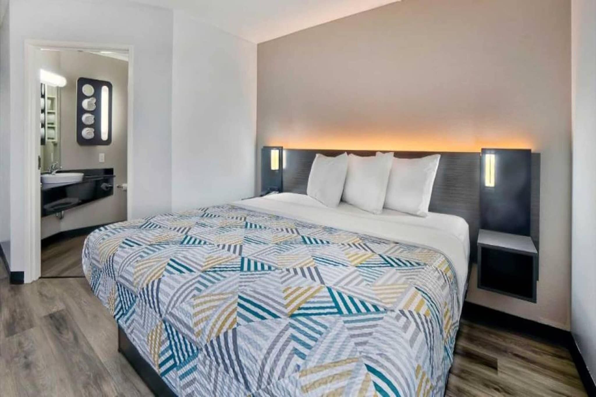 A modern hotel room with a large bed, a bedside table, a nightstand, a built-in shelf, and a bathroom visible through an open door.