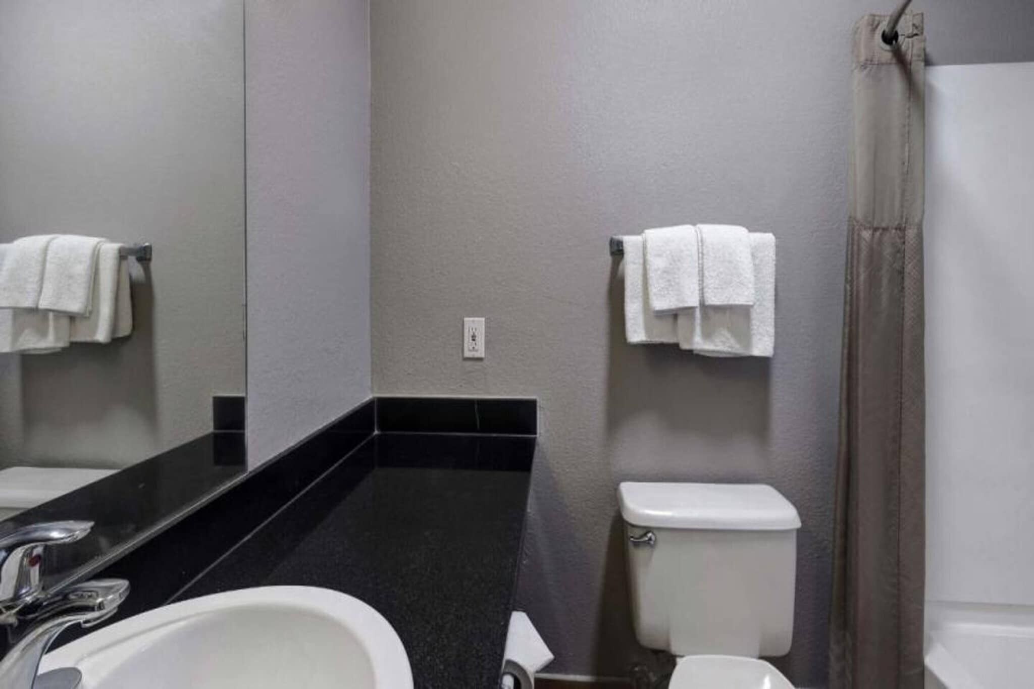 A bathroom with a black countertop, white sink, and toilet. There are towels hanging on the wall.