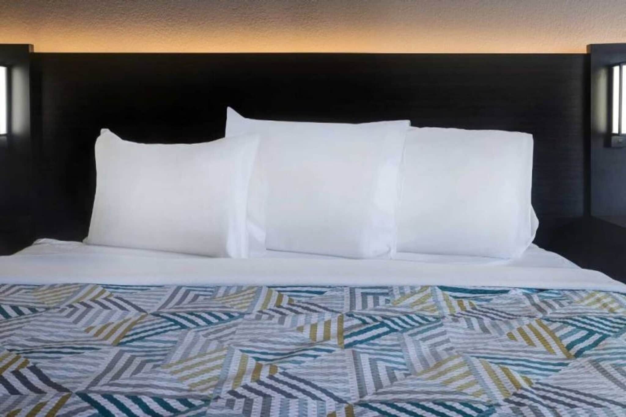 Una cama con ropa de cama blanca, un plaid estampado, un cabecero oscuro y luminarias fijadas a la pared.