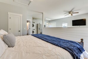 Ein Schlafzimmer mit einem Bett, einem Deckenventilator, einem an der Wand befestigten Fernseher und einem Waschbereich, der durch eine geöffnete Tür sichtbar ist.