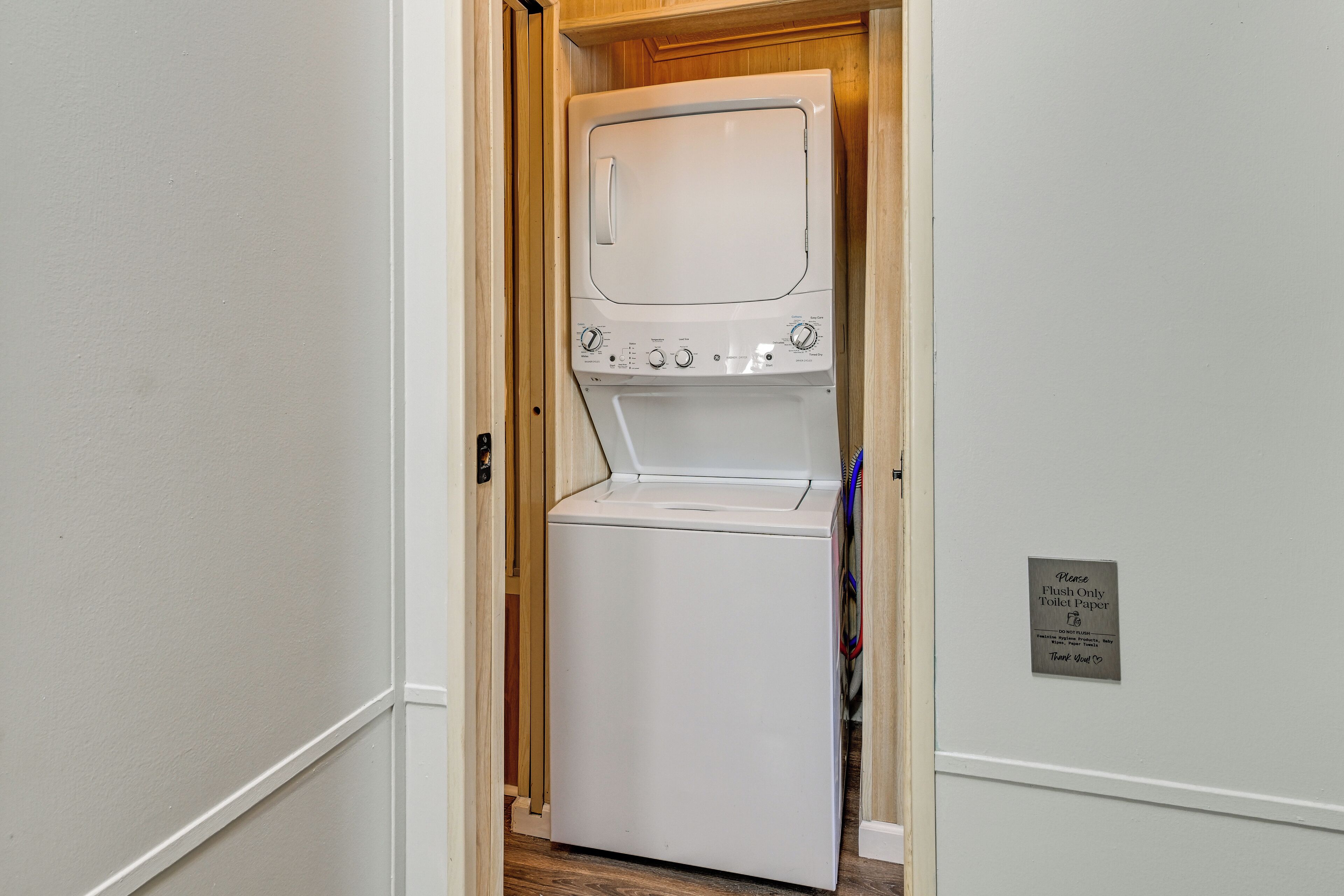 A laundry room with a white washer and dryer, a wooden door, and a sign instructing to flush only toilet paper.