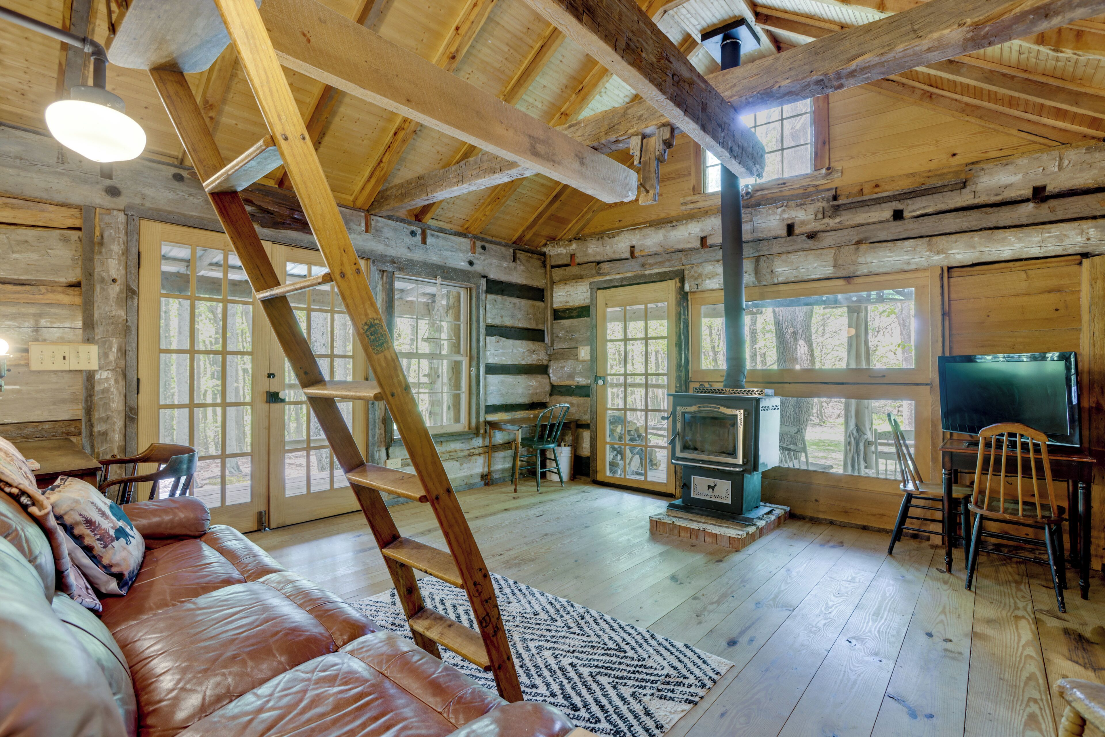 A log cabin interior with a wooden staircase, a leather couch, a fireplace, and large windows.