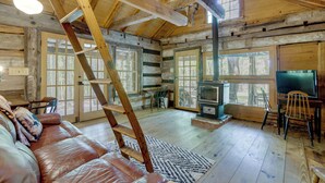 A log cabin interior with a wooden staircase, a leather couch, a fireplace, and large windows. - Mountain Tree House w/ Views in Maysville (Maysville)