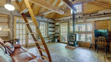 A log cabin interior with a wooden staircase, a leather couch, a fireplace, and large windows.