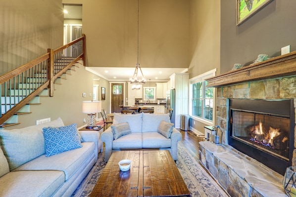 A spacious living room with a fireplace, a staircase, and a kitchen in the background.