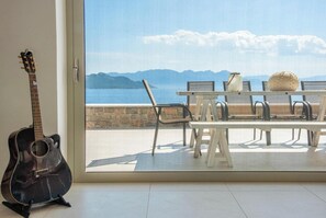 Eine Gitarre, ein Tisch mit Stühlen und Blick auf das Meer und die Berge durch eine Glastür.