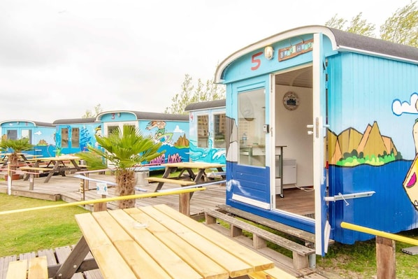 Une rangée de maisons mobiles bleues, chacune avec une porte numérotée, des tables de pique-nique et un palmier.