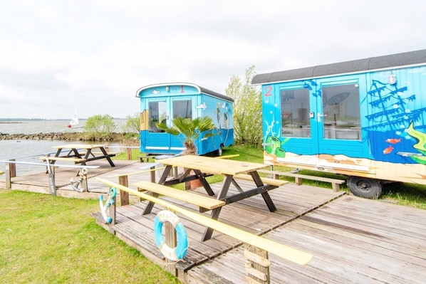 Twee blauwe stacaravans met picknicktafels en een reddingsboei, afgemeerd aan een water.