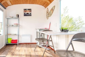 Een kamer met een houten plafond, een witte muur met een afbeelding, een plank met spullen, een tafeltje, een stoel en een raam met uitzicht.