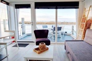 Una habitación con sofá, una mesa, una silla, un ventanal con vistas al agua y un balcón con mobiliario exterior.