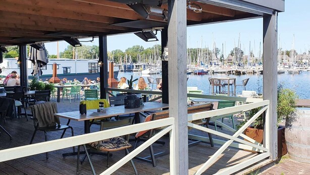 An outdoor dining area with tables and chairs, overlooking a marina with boats. - Cozy Tiny Wagon by the Marina (Naarden)