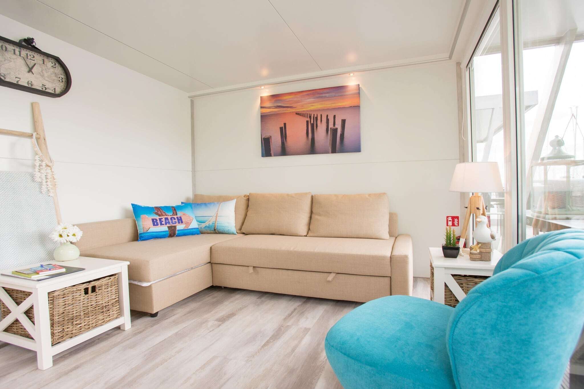 Un salon moderne avec un canapé d’angle beige, un fauteuil bleu, une horloge murale et un tableau représentant un coucher de soleil.