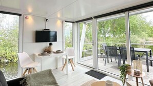 Un intérieur moderne doté d’un téléviseur à écran plat, d’un coin repas avec des chaises et une table, ainsi que de grandes portes-fenêtres coulissantes donnant sur une terrasse extérieure.