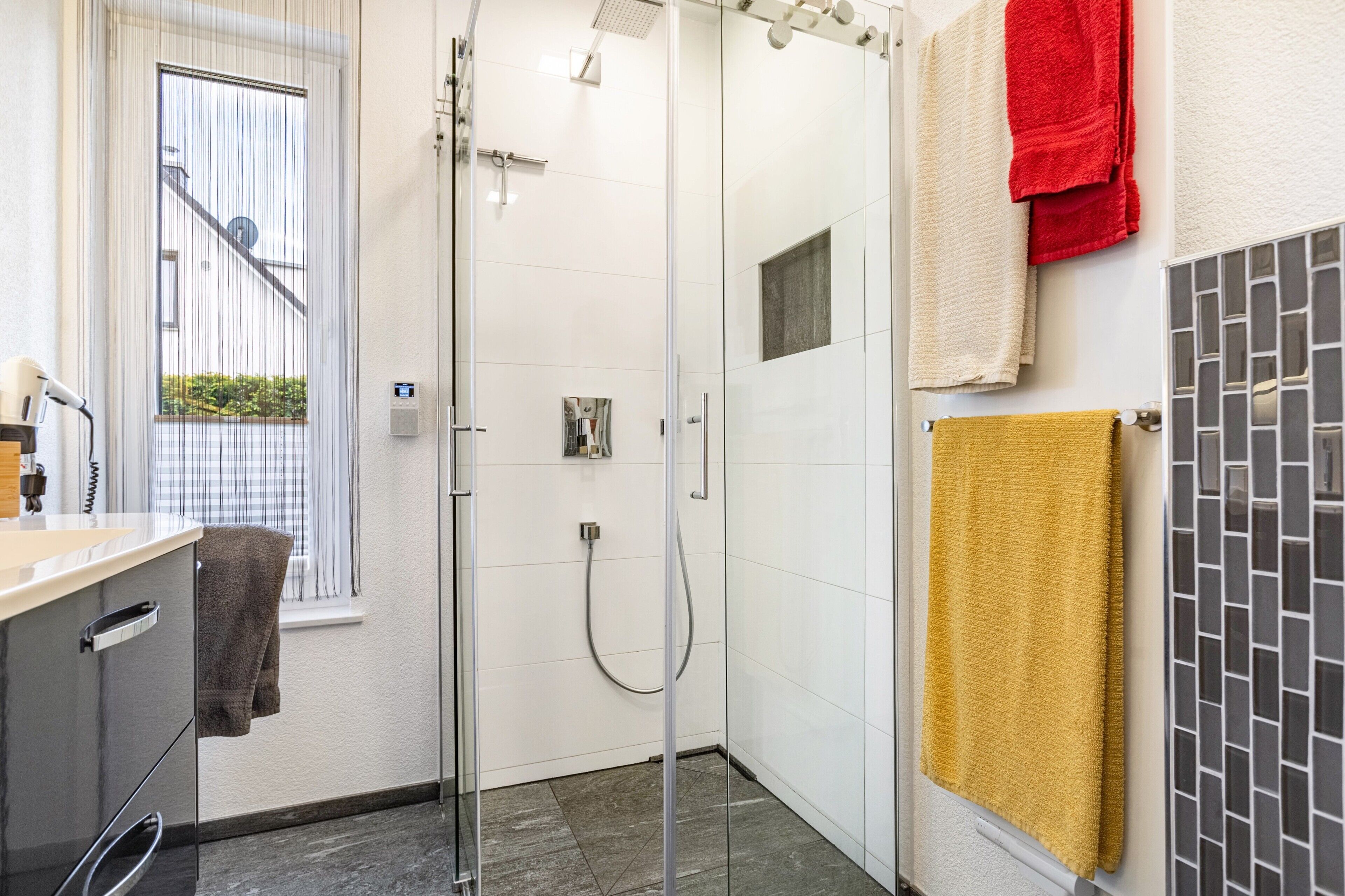 A modern bathroom with a glass shower enclosure, a white tiled wall, and a window with a view.