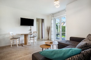 A modern living room with a flat-screen TV, a dining table, two chairs, a sofa, and a small coffee table. - Apartment 'Für 2 Personen 35 M²' with Private Terrace, Private Garden and Wi-Fi (Koserow)
